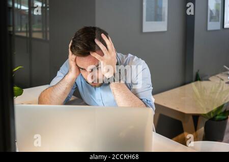 Uomo d'affari stanco con la testa nelle mani che guarda il laptop scrivania in ufficio creativo Foto Stock