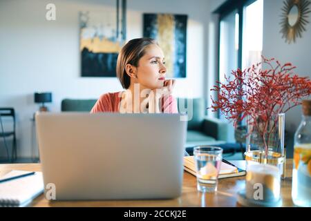 Giovane donna premurosa con un computer portatile sulla scrivania che guarda lontano seduto a casa Foto Stock