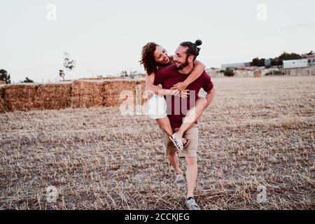 Uomo sorridente che dà alla donna piggyback cavalcare contro la paglia in campo Foto Stock