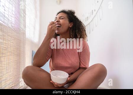 Donna allegra che mangia crema e fragole mentre si siede a muro a casa Foto Stock