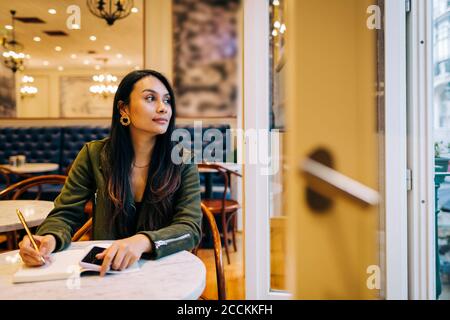 Giovane donna che guarda fuori mentre scrive nel libro al caffè Foto Stock