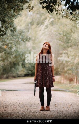 Sorridente giovane donna che tiene clarinetto mentre si guarda in su all'albero e in piedi sul sentiero nella foresta Foto Stock