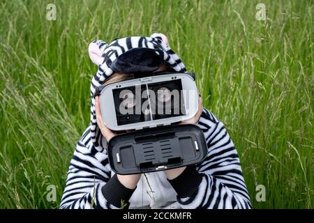 Ragazzo che indossa un costume zebra utilizzando un simulatore VR mentre si siede erba Foto Stock