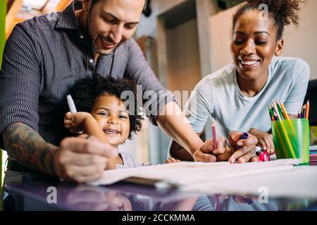 Sorridente genitori che guardano la figlia allegra che tiene la penna della punta del feltro a tavola Foto Stock