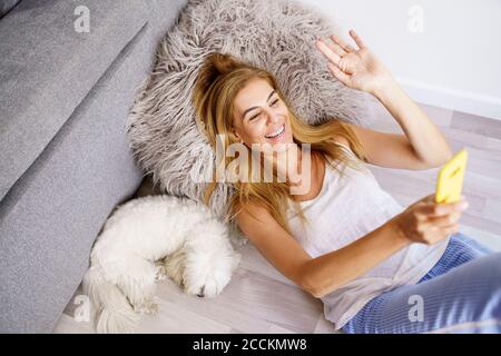 Donna sorridente che agita la mano mentre videochiama tramite smartphone a casa Foto Stock