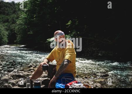 Escursionista con barba completa e felpa con cappuccio gialla durante la pausa, cucinando il tè sul lungofiume Foto Stock