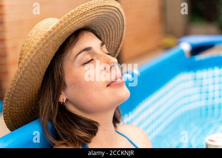 Giovane donna che indossa un cappello da sole mentre si rilassa nel nuoto gonfiabile piscina nel cortile Foto Stock