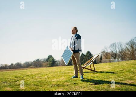 Uomo d'affari che guarda via mentre tiene il pannello solare dalla sedia sopra erba al parco contro il cielo limpido durante la giornata di sole Foto Stock