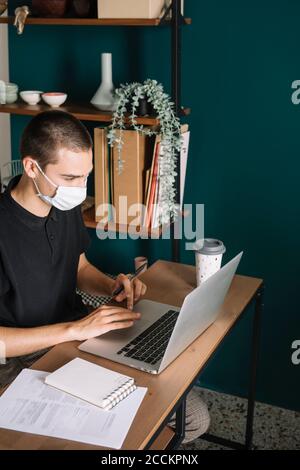 Giovane uomo che indossa una maschera protettiva utilizzando un computer portatile sulla scrivania casa Foto Stock