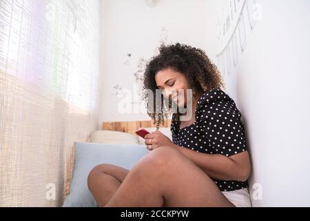 Sorridente giovane donna con capelli ricci utilizzando smartphone mentre seduto contro il muro a casa Foto Stock