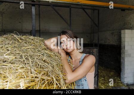 Giovane donna con gli occhi chiusi che pende sulla paglia in un fienile in una fattoria Foto Stock