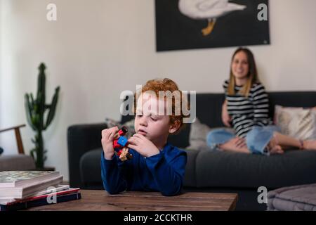 Madre che si rilassa sul divano mentre il figlio gioca con il robot giocattolo sul tavolo a casa Foto Stock