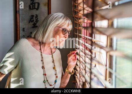 Donna anziana che indossa occhiali da sole guardando attraverso la finestra a casa Foto Stock