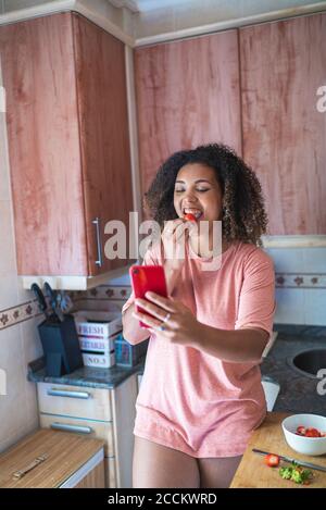 Giovane donna allegra che mangia la fragola mentre videochiama su Smart telefono in cucina Foto Stock