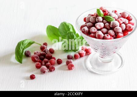 Mirtilli congelati in un recipiente di vetro. Mirtilli rossi e foglie verdi di basilico sulla tavola bianca. Foto Stock