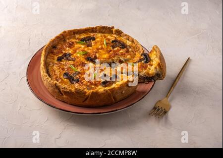 Quiche tradizionali francesi a torta con funghi e formaggio con un pezzo tagliato su un piatto, accanto a una forchetta, su uno sfondo chiaro testurizzato, fuoco selettivo, primo piano Foto Stock