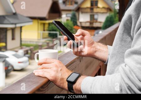 Uomo in orologio intelligente bere caffè o tè sul balcone sullo sfondo di ville o pensioni, primo piano mans Hand utilizzando il suo smartphone. Concetto di Foto Stock