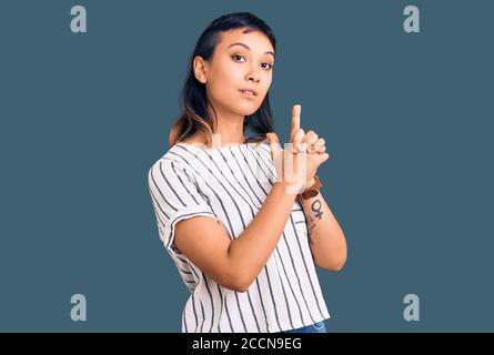 Giovane donna che indossa abiti casual che reggono una pistola simbolica con gesto della mano, giocando uccidendo armi di tiro, faccia arrabbiata Foto Stock