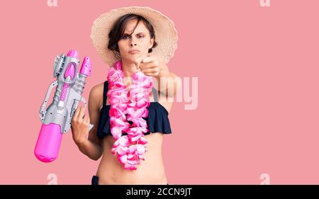 Bella giovane donna con capelli corti indossando bikini e hawaian lei tenendo pistola ad acqua che punta con il dito alla macchina fotografica e a voi, sicuro gestu Foto Stock