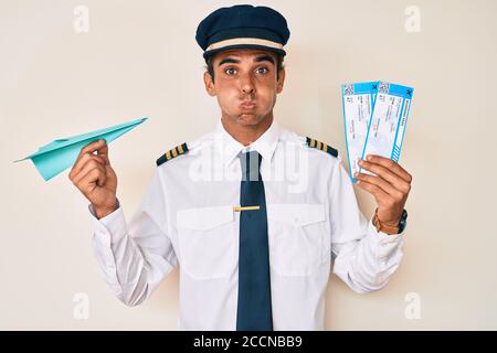 Giovane uomo ispanico che indossa un aereo pilota uniforme tenendo carta aereo e carta d'imbarco guance soffiato con viso divertente. Bocca gonfia con aria, ca Foto Stock