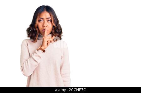 Giovane bella donna mista da corsa con maglione invernale a dolcevita che si chieda di essere tranquilla con le dita sulle labbra. Silenzio e concetto segreto. Foto Stock