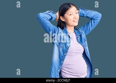 Giovane bella donna cinese incinta aspettandosi il bambino che si distende e si distende, braccia e mani dietro la testa e il collo sorridendo felice Foto Stock