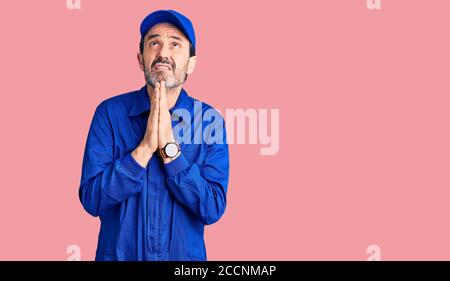 Bell'uomo di mezza età che indossa uniforme meccanica che prega e che prega con le mani insieme con espressione di speranza sul viso molto emotivo e preoccupato Foto Stock