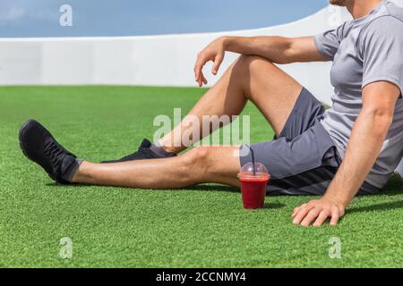 Smoothie smoothie uomo sportivo rilassante godendo della prima colazione del mattino dopo l'allenamento seduto su erba all'aperto a casa o palestra fitness. Atleta che beve frutta rossa Foto Stock