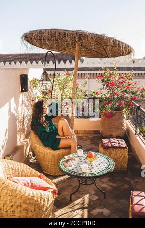 Donna che gusterà la colazione sulla splendida terrazza dell'hotel lounge nel centro di Marrakech all'alba. Design marocchino con elementi vintage - lanterna in metallo, sedia in vimini, tavolo rotondo e ombrello. Foto Stock