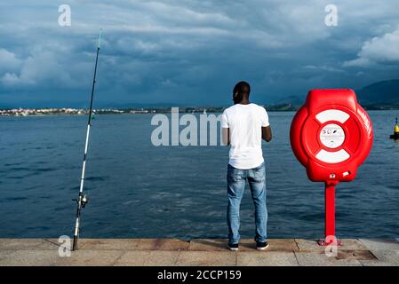 Vista posteriore di un giovane uomo nero che indossa una T-shirt bianca. Uomo nero che ascolta la musica mentre pesca in Paseo Maritimo, Santander, Spagna Foto Stock
