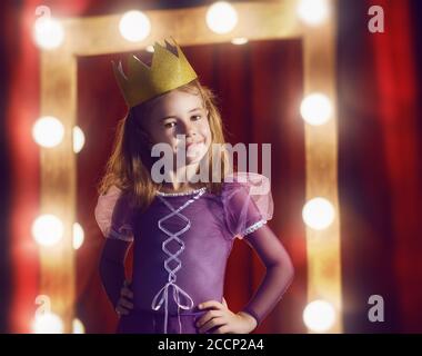 Carino attrice piccola. Bambina in costume principessa sullo sfondo di scene teatrali e specchi. Foto Stock