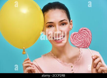 Giovane donna divertente sullo sfondo di una parete blu brillante. Bella ragazza si sta divertendo con palloncino e lollipop. Colori giallo, rosa e turchese. Foto Stock