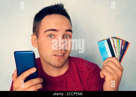 Un giovane che tiene uno smartphone in una mano nell'altra un sacco di carte di debito diverse su uno sfondo blu. Scelta della carta bancaria per il pagamento in Foto Stock