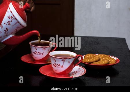 Versare il tè in una tazza di ceramica su un tavolo di legno con un po' di biscotto sul piatto. Messa a fuoco parziale. Foto Stock