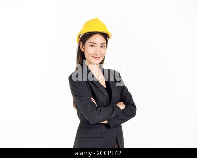 Giovane donna d'affari asiatica sorridente indossando tuta nera e casco di sicurezza giallo in piedi con le braccia piegate. Ritratto di ingegnere femminile guardando confi Foto Stock