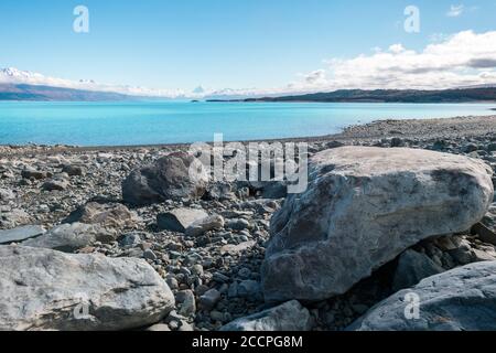 Lago Pukaki di fronte alle montagne innevate delle alpi meridionali in Nuova Zelanda con il Monte Cook che torreggia soprattutto. Enormi rocce in primo piano Foto Stock