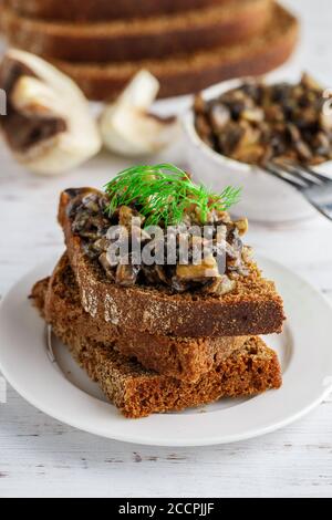 Pane di segale integrale con funghi fritti con cipolla, aglio, panna e aneto su un piatto bianco su un tavolo di legno chiaro. L'antipasto di funghi. Foto Stock