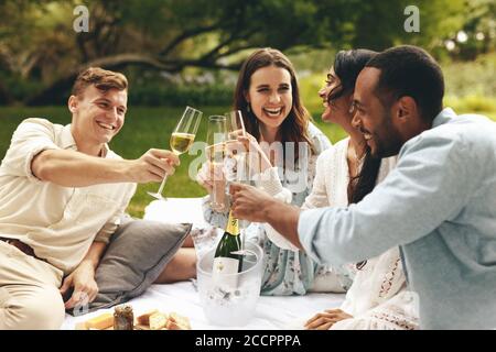 Amici multietnici che sorseggiano bicchieri di champagne mentre si siedono al parco. Due coppie che si godono un picnic di lusso in campagna. Foto Stock