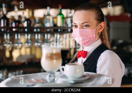 Una cameriera femminile di aspetto europeo in una maschera medica serve caffè latte. Foto Stock
