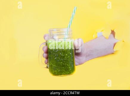 Frullato di erbe fresche in un vaso di vetro è tenuto con la mano di un uomo in un foro su un giallo sfondo Foto Stock