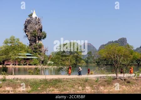 Monaci che camminano di fila di fronte alla pagoda Kyaut Ka Latt, monastero e roccia sacra circondato da un lago. Indossare abiti arancioni. HPa An, Myanmar Foto Stock