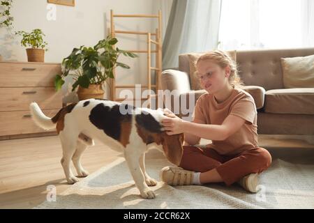 Ritratto di una bella ragazza dai capelli rossi che gioca con il cane mentre si siede sul pavimento in un accogliente interno di casa illuminato dalla luce del sole, spazio di copia Foto Stock