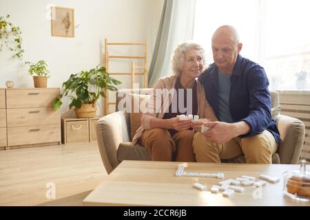 Ritratto dai toni caldi della coppia senior felice che tiene i pezzi domino mentre si giocano i giochi da tavolo a casa, spazio di copia Foto Stock