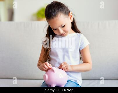 Bambino ragazza mettere moneta in Piggybank con risparmi a casa Foto Stock