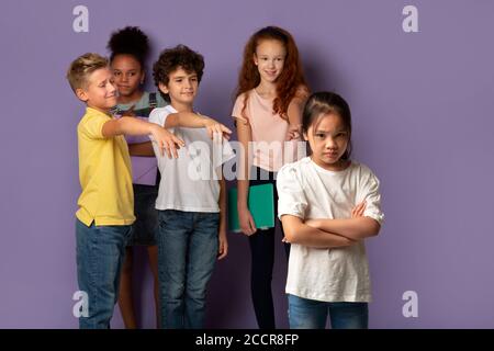 Smetta di bullonare nelle scuole. Infelice ragazza asiatica e i suoi compagni di classe crudeli che la puntano su uno sfondo lilla Foto Stock