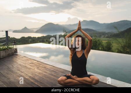 Donna che medita da sola a bordo piscina infinity con splendida vista sull'oceano e sulle montagne al mattino. Stile di vita sano, concetto spirituale ed emotivo. Risveglio, armonia con la natura. Foto Stock