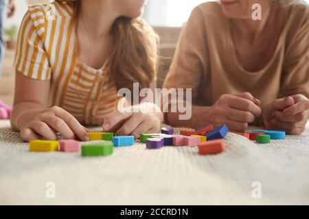 Primo piano dai toni caldi di una ragazza irriconoscibile che gioca con blocchi colorati mentre giace sul pavimento con la nonna in un accogliente interno, spazio di copia Foto Stock