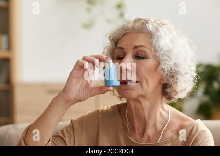 Testa e spalle dai toni caldi ritratto di donna anziana uso di inalatore per asma o problemi respiratori in ambiente domestico Foto Stock