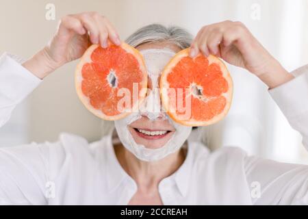 Primo piano di bella donna anziana con maschera facciale sul viso che tiene fette di pompelmo fresco che copre gli occhi, sullo sfondo interno casa Foto Stock