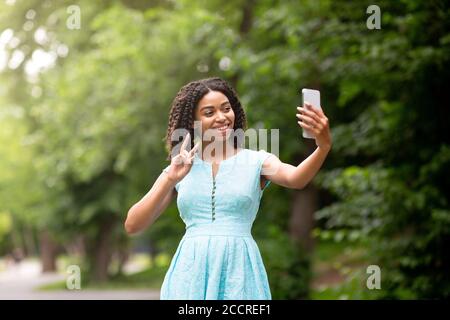 Bella ragazza afroamericana prendendo selfie o comunicando in linea sopra telefono cellulare al parco Foto Stock